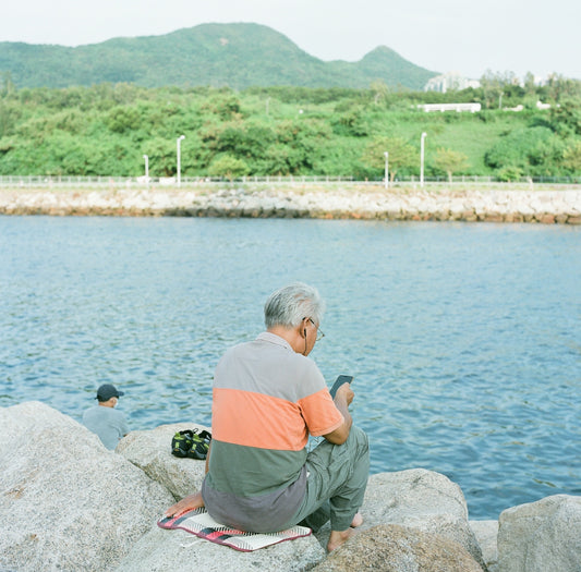 man in orange t-shirt and gray pants sitting on gray rock near body of water
