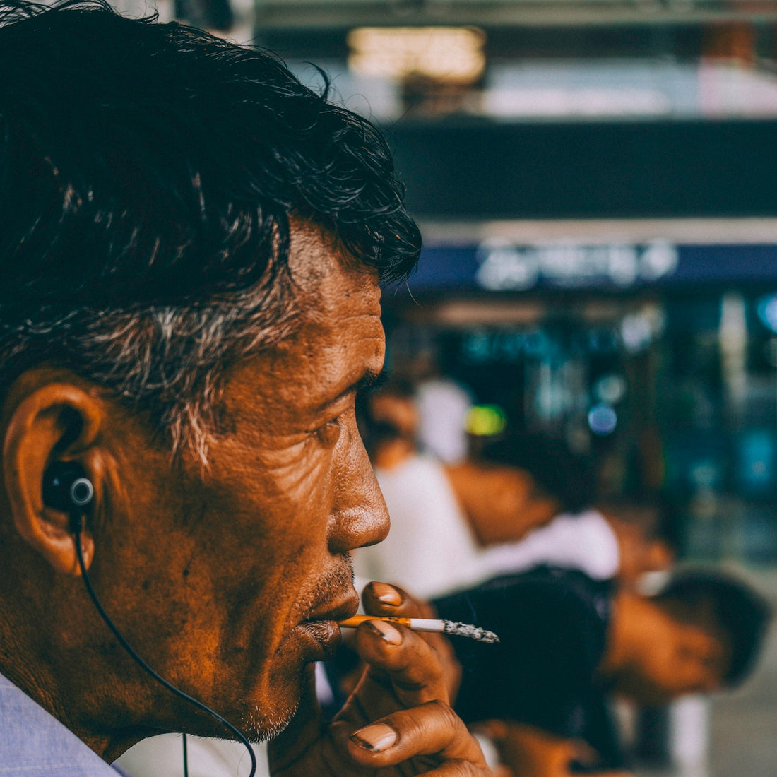 A man with dark hair wearing earbuds and smoking a cigarette in profile