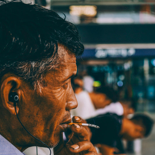 A man with dark hair wearing earbuds and smoking a cigarette in profile